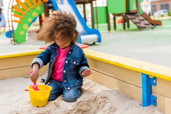 Niño afroamericano rizado jugando con cuchara de plástico y cubo en caja de arena en el patio de recreo - foto de stock