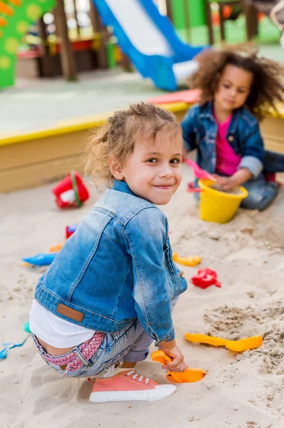 Selective focus of smiling little child sitting in sandbox with plastic scoop while her friend sitting behind at playground — Stock Photo