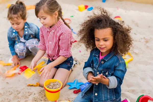 Foco seletivo da criança afro-americana encaracolada brincando de amigos sagazes em sandbox no playground — Fotografia de Stock