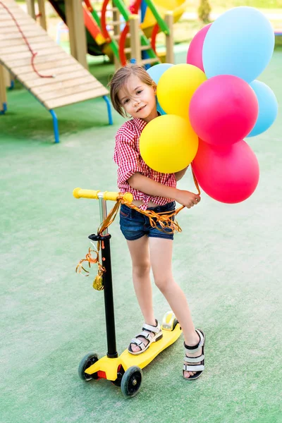 Enfoque selectivo de niño pequeño montando en patinete scooter con globos de colores en el patio de recreo - foto de stock