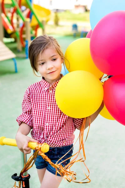 Очаровательный маленький ребенок едет на самокате с красочными воздушными шарами на детской площадке — стоковое фото