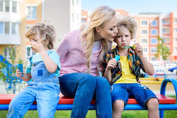 Madre sentada entre dos adorables hijitos mientras usan sopladores de burbujas en el banco - foto de stock
