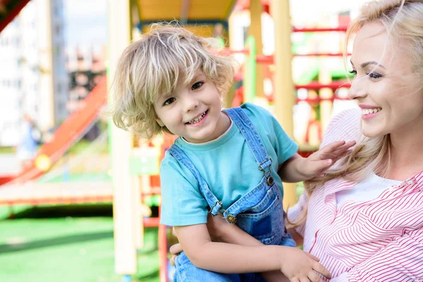 Selective focus of smiling mother embracing and holding little son at playground — Stock Photo