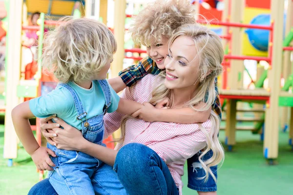Mãe feliz abraçando com dois filhos adoráveis brincalhão no parque infantil — Fotografia de Stock
