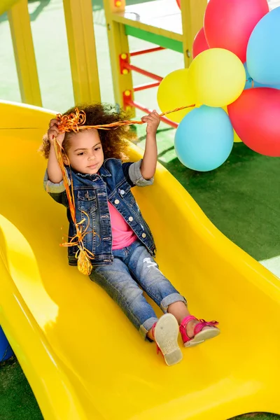 Curly african american little child holding colorful balloons and sliding down from hill at playground — Stock Photo
