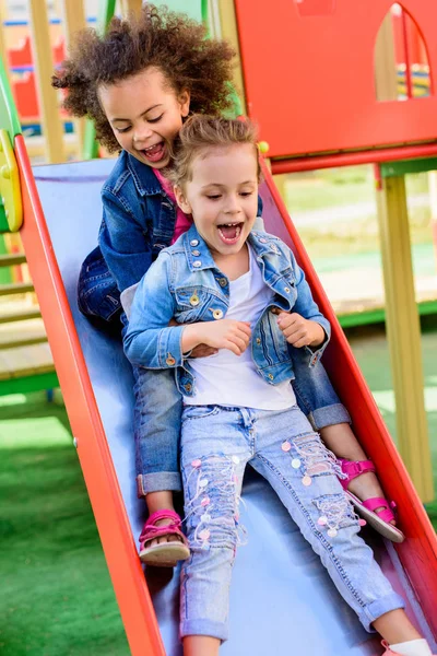 Due eccitati urlando bambini multietnici che scivolano giù dalla collina al parco giochi — Foto stock