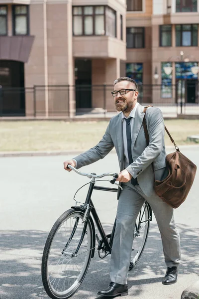 Hombre de negocios guapo en traje gris y gafas caminando con bicicleta en la calle en la ciudad - foto de stock