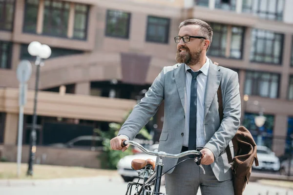 Homem de negócios bonito andando com bicicleta na rua na cidade e olhando para longe — Stock Photo