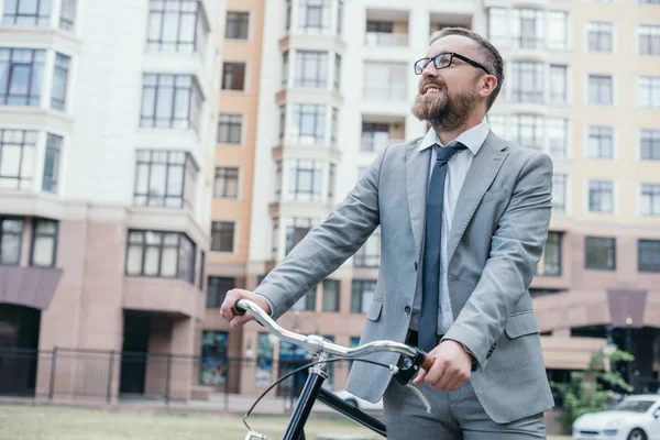 Handsome businessman walking with bike on street in city and looking up — Stock Photo