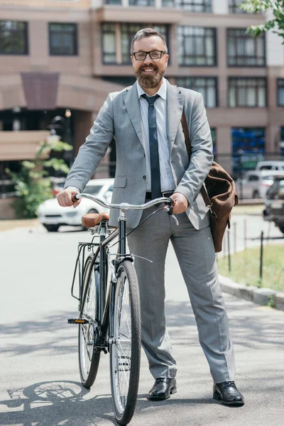 Homem de negócios bonito feliz de pé com bicicleta e olhando para a câmera na rua na cidade — Fotografia de Stock