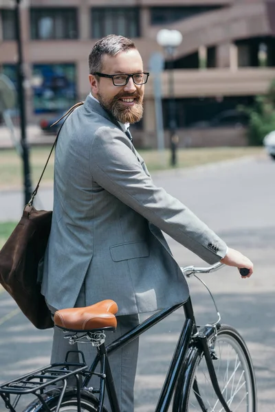 Smiling handsome businessman standing with bike and looking at camera on street in city — Stock Photo