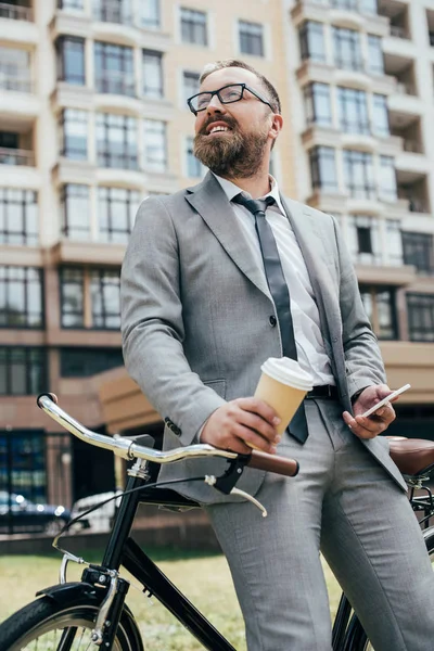 Handsome bearded businessman with coffee and smartphone leaning on bicycle — Stock Photo