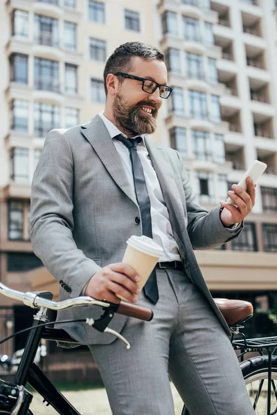 Homme d'affaires barbu avec tasse de café jetable et smartphone appuyé sur le vélo — Photo de stock