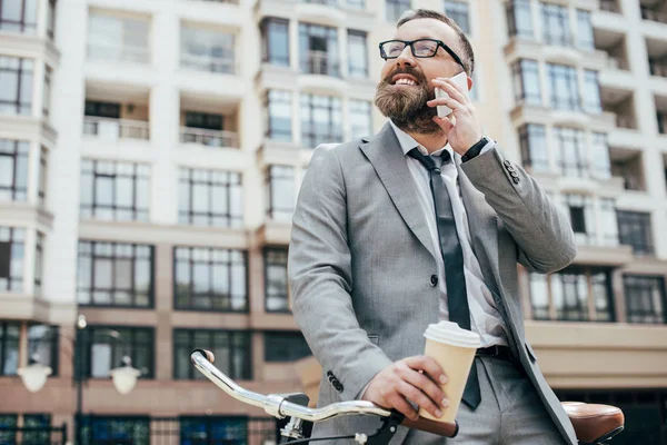 Homme d'affaires barbu avec vélo tenant une tasse de café jetable et parlant sur smartphone — Photo de stock