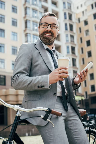 Hombre de negocios barbudo con bicicleta que sostiene la taza desechable de café y teléfono inteligente - foto de stock