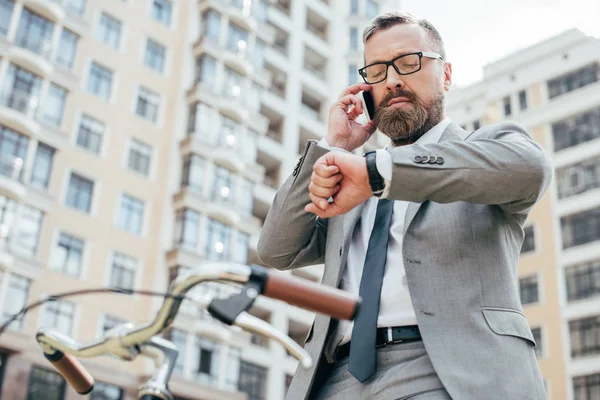 Der bärtige Geschäftsmann spricht auf dem Smartphone und schaut auf die Uhr, während er auf dem Fahrrad sitzt. — Stockfoto