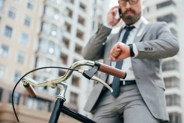 Enfoque selectivo de hombre de negocios hablando en el teléfono inteligente y mirando reloj de pulsera mientras está sentado en bicicleta - foto de stock