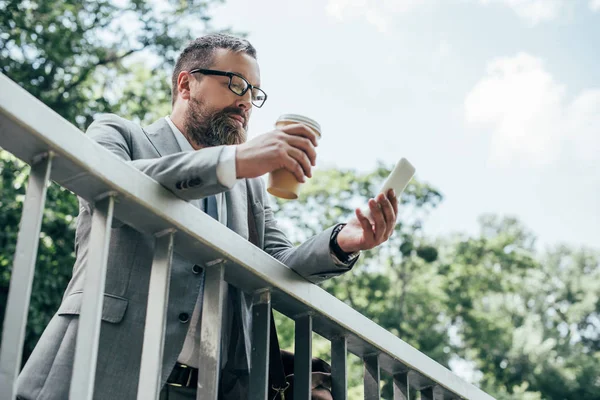 Homem de negócios barbudo com xícara descartável de café usando smartphone — Fotografia de Stock