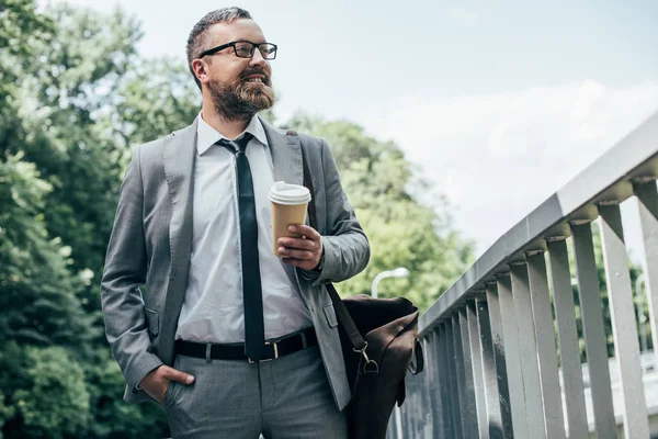 Hombre de negocios barbudo en traje con bolsa de cuero y café para ir caminando por la ciudad - foto de stock