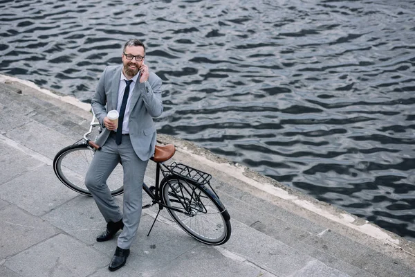 Hombre de negocios con café para ir a hablar en smartphone y de pie con bicicleta en muelle — Stock Photo