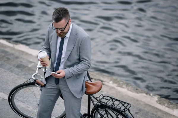 Hombre de negocios con café para llevar y smartphone de pie con bicicleta en muelle — Stock Photo