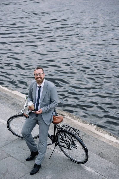 Businessman with coffee to go and smartphone standing with bicycle on quay — Stock Photo