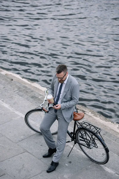 Businessman with coffee to go using smartphone and standing with bike on quay — Stock Photo