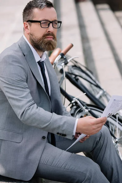 Pensive bearded businessman reading newspaper on stairs with bicycle — Stock Photo