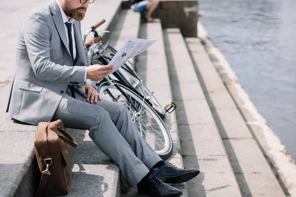 Businessman in grey suit reading newspaper on stairs on quay with bicycle and leather bag — Stock Photo