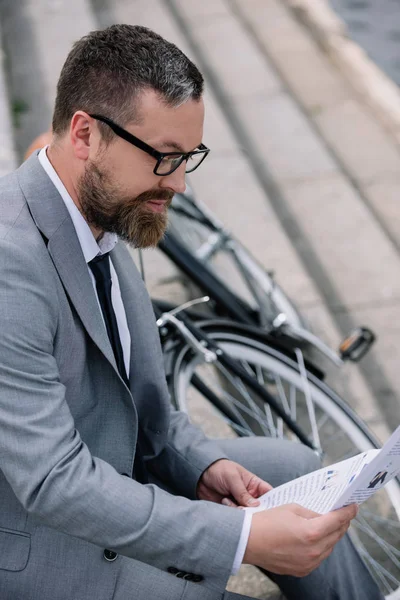 Handsome bearded businessman reading newspaper on stairs with bicycle — Stock Photo