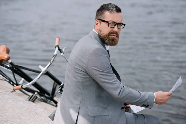 Bärtiger Geschäftsmann mit Zeitung sitzt mit Fahrrad am Kai — Stockfoto