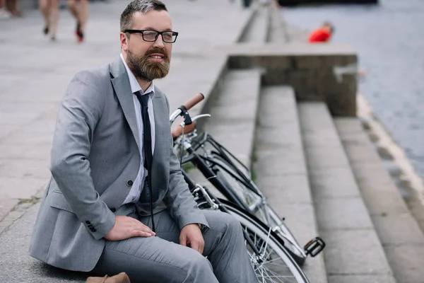 Handsome bearded businessman in grey suit sitting on stairs on quay with bicycle — Stock Photo