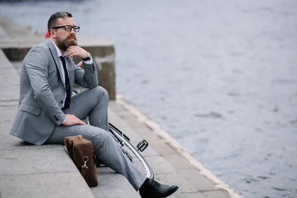 Handsome thoughtful businessman in grey suit sitting on stairs on quay — Stock Photo