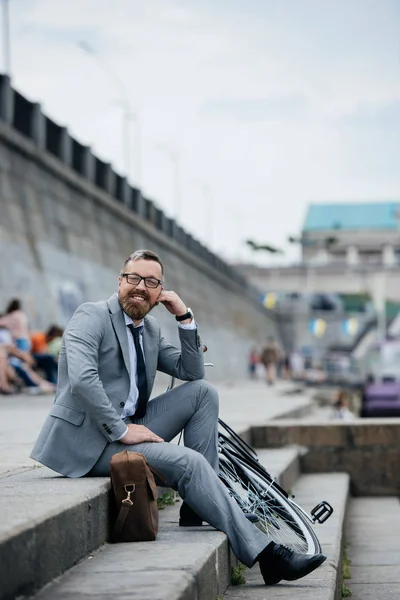 Uomo d'affari sorridente in abito grigio seduto sulle scale con bicicletta — Foto stock
