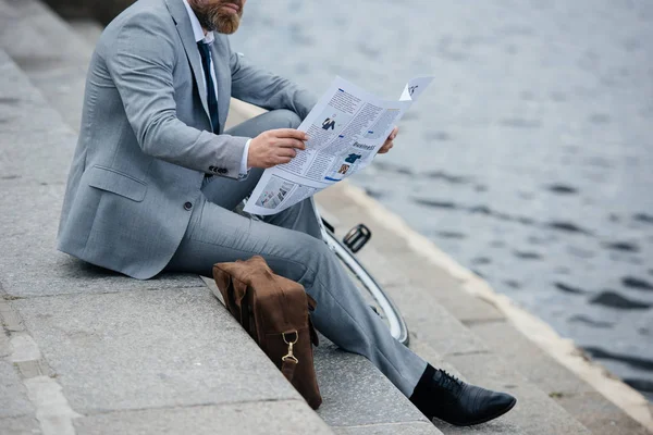 Vista recortada del hombre de negocios barbudo en traje gris leyendo periódico en muelle - foto de stock