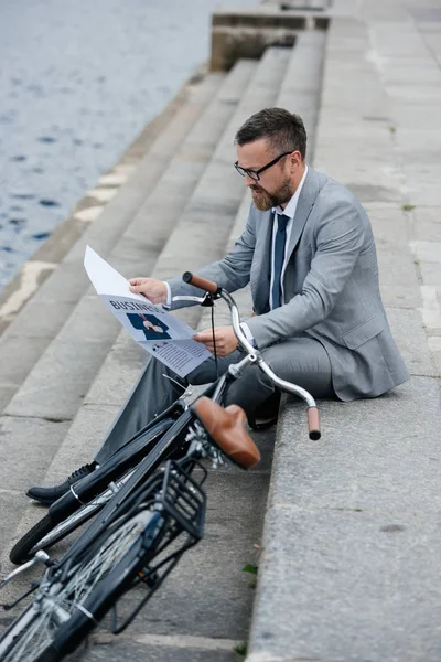 Bearded businessman in grey suit reading newspaper on quay with bike — Stock Photo