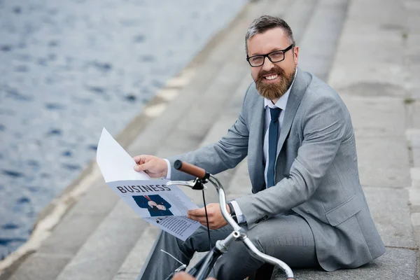 Bonito sorridente empresário em cinza terno ler jornal no cais com bicicleta — Fotografia de Stock