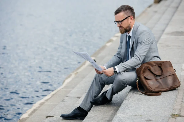 Handsome businessman in grey suit with leather bag reading newspaper on quay — Stock Photo