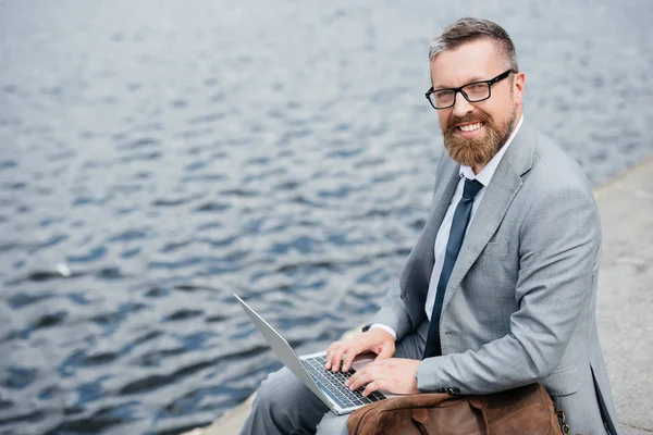 Handsome businessman using laptop and sitting on quay with leather bag — Stock Photo