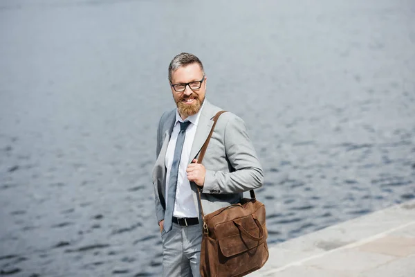 Handsome businessman with leather bag walking on quay near river — Stock Photo