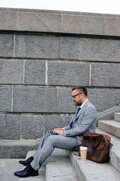 Hombre de negocios barbudo con bolsa y taza de café desechable utilizando el ordenador portátil y sentado en las escaleras - foto de stock