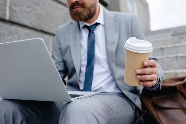 Vista recortada de hombre de negocios utilizando el ordenador portátil y la celebración de café para ir - foto de stock