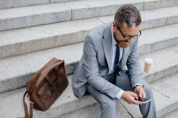 Homme d'affaires utilisant smartphone et assis sur les escaliers avec sac en cuir et café — Photo de stock