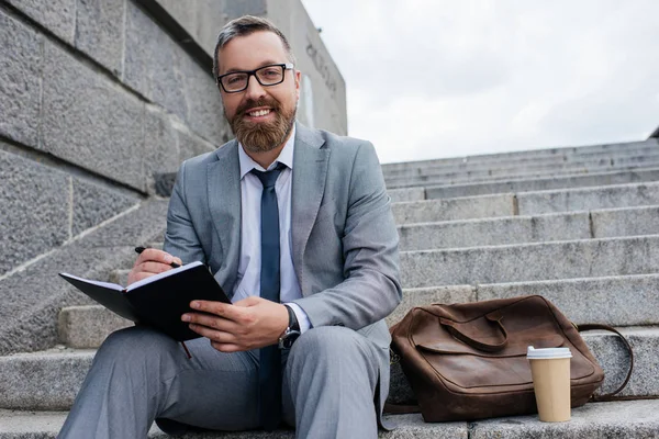 Businessman with leather bag and coffee to go writing in diary and sitting on stairs — Stock Photo