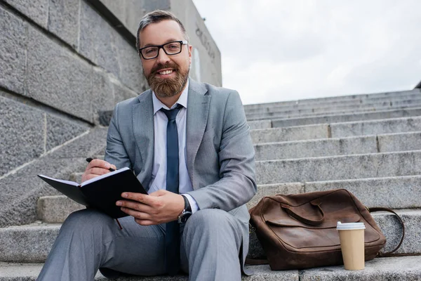 Homme d'affaires avec sac en cuir et café pour aller écrire dans le journal et assis sur les escaliers — Photo de stock