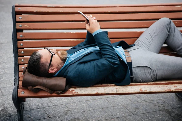 Homem de negócios usando smartphone deitado no banco na cidade — Fotografia de Stock