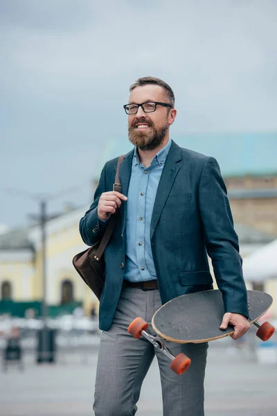 Hombre barbudo con estilo con bolso de cuero y longboard caminando en la ciudad - foto de stock