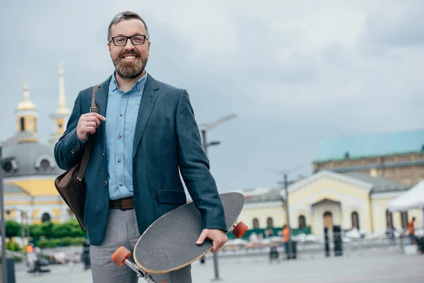 Smiling bearded man with leather bag and longboard walking in city — Stock Photo