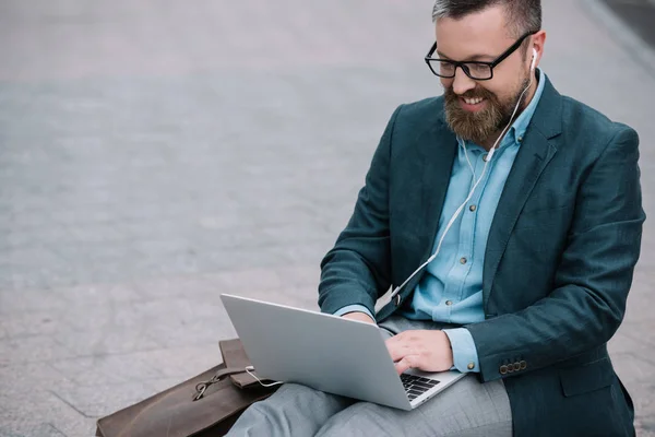 Hombre barbudo con estilo utilizando el ordenador portátil con auriculares y sentado en la ciudad - foto de stock