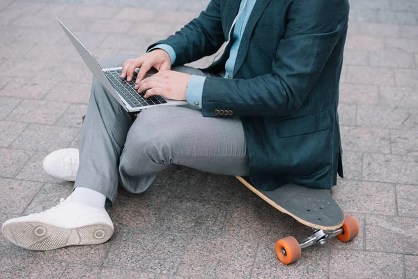 Cropped view of man using laptop and sitting on skateboard — Stock Photo